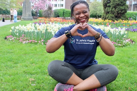 A supporter wears her navy blue St John's Hospice t-shirt, running leggings and trainers. She sits cross legged on the grass in the park with flowers behind her.  She is holding her hands to make the gesture of a heart.