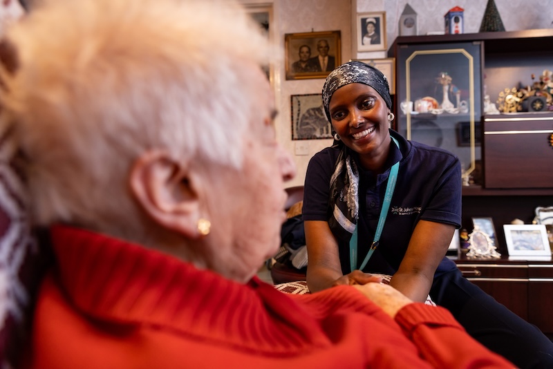 A Hospice@Home Healthcare Assistant is smiling and holding a patient's hand whose face we cannot see. They are talking in the patient's own living room at home.
