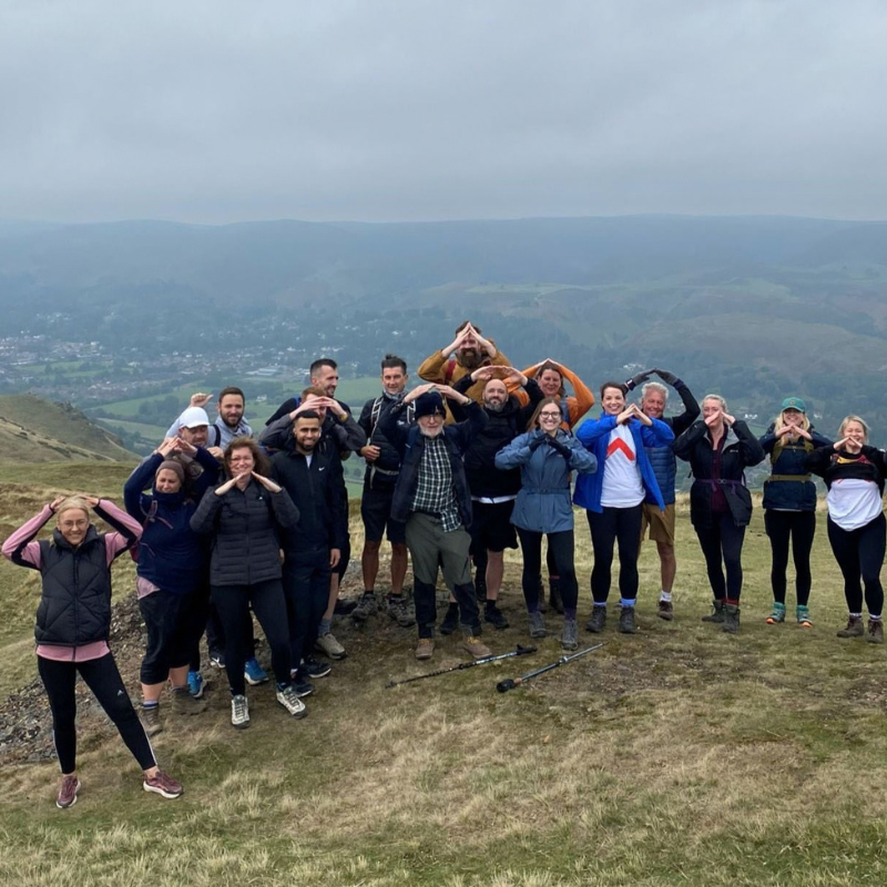 A group of people, mixed in gender, age and ethnicity, stood on a peak of the shropshire hills, using their arms above their head to make the shelter logo