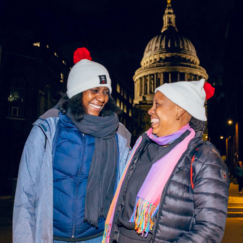 Two women wrapped up in warm winter jackets, scarves, and matching beanie hats topped with red pom-poms, smiling together at night with the lit-up dome of St Paul's Cathedral visible behind them.