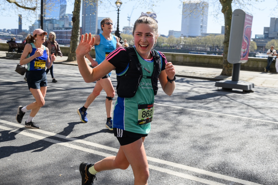 A young blonde woman running along the thames. She is waving to the camera as she runs past.