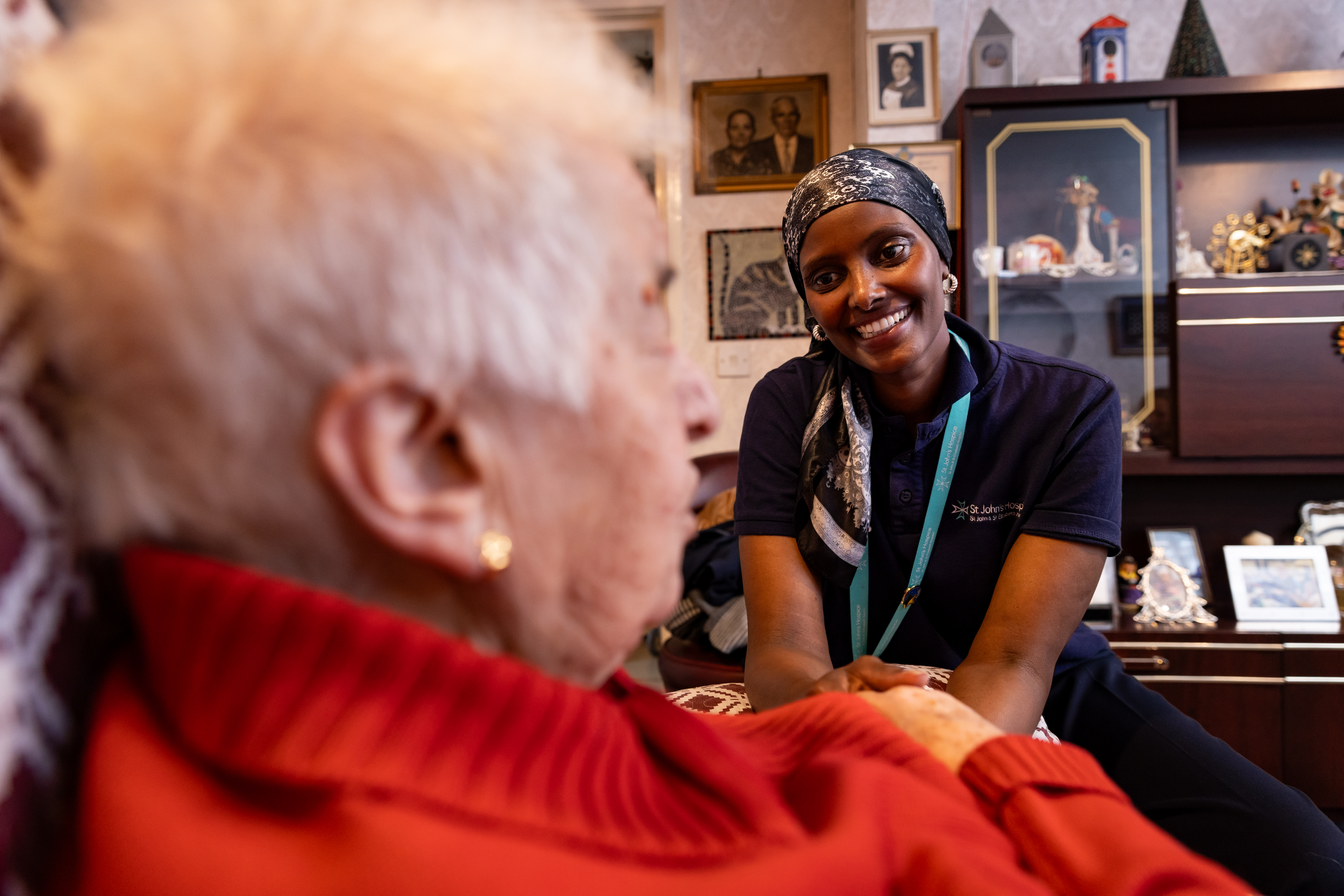 A Hospice@Home Healthcare Assistant is smiling and holding a patient's hand whose face we cannot see. They are talking in the patient's own living room at home.